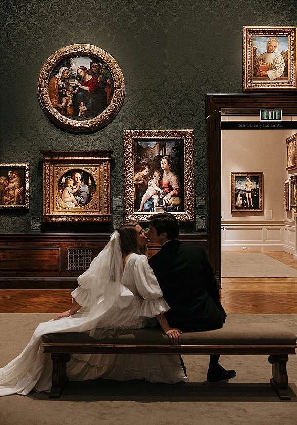 Wedding kiss portrait of bride and groom kissing on a bench, her long veil draped over his black suit in an art gallery with framed paintings
