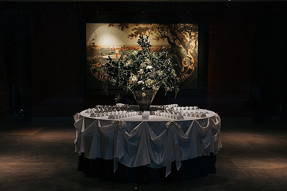 Escort card table with wedding escort cards beneath a large white-flower and greenery centerpiece in a glass vase, spotlighted in a dark room