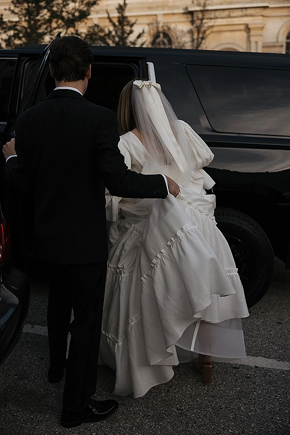 Wedding exit as groom in black tuxedo helps bride in puff-sleeve gown and bow veil into a black SUV on a tree-lined street