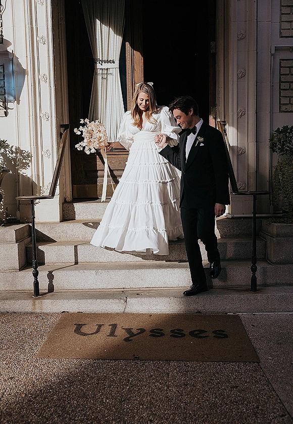 Couple portrait of newlyweds walking down steps, holding hands with a bridal bouquet and ribbon wrap at a stone doorway entrance
