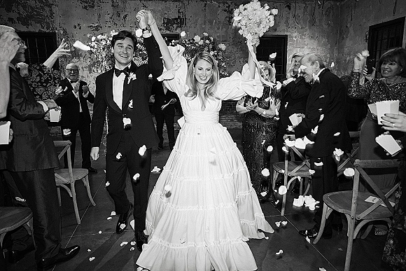 Wedding recessional as newlyweds walking aisle, hands raised while guests toss flower petals in an indoor space with a floral arch backdrop