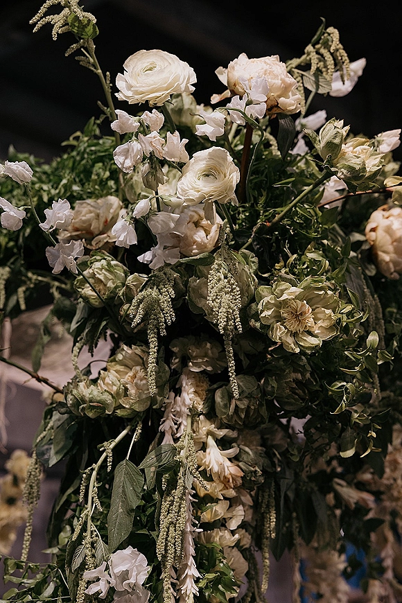 Wedding floral arrangement of white wedding flowers with cream roses, ranunculus, and cascading greenery against a dark blurred reception backdrop