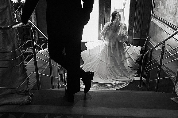 Bridal portrait in a black and white wedding portrait style of bride walking down stairs, cathedral veil and long train, groom in tuxedo behind in hallway