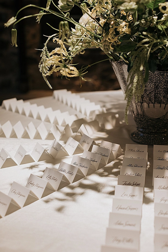 Wedding place cards with calligraphy place cards folded on a white tablecloth, beside a floral centerpiece in a wire vase with sunlight shadows