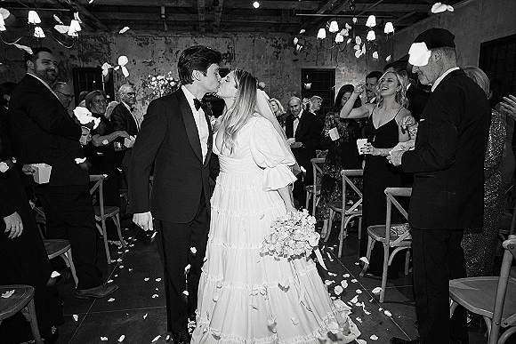 Wedding recessional as bride and groom kiss in the aisle while guests cheer and toss rose petals, bouquet and veil under chandeliers