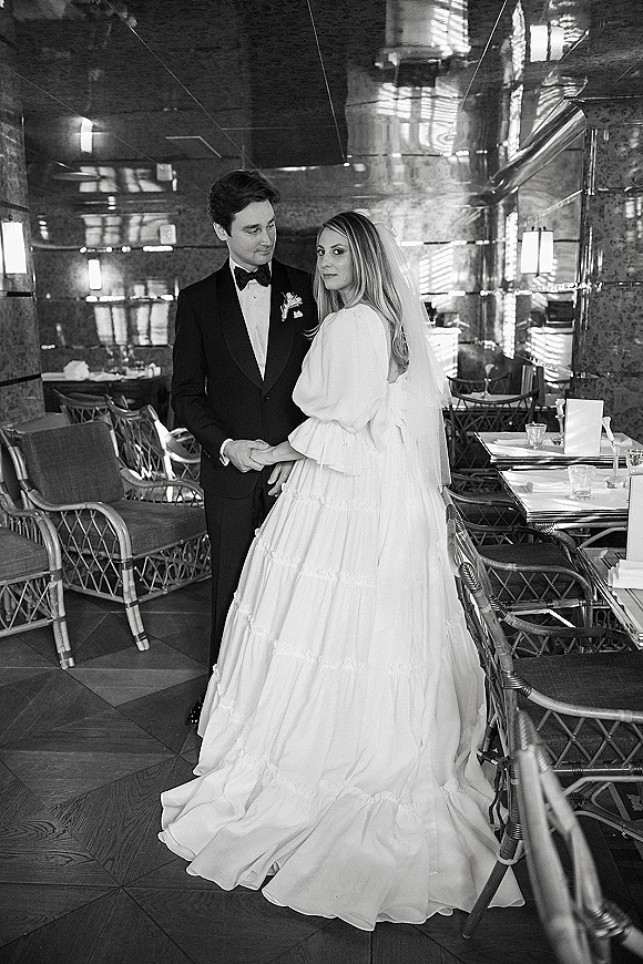 Couple portrait in a black and white wedding photo, bride in veil and long-sleeve gown holding hands with groom in tuxedo inside a mirrored restaurant
