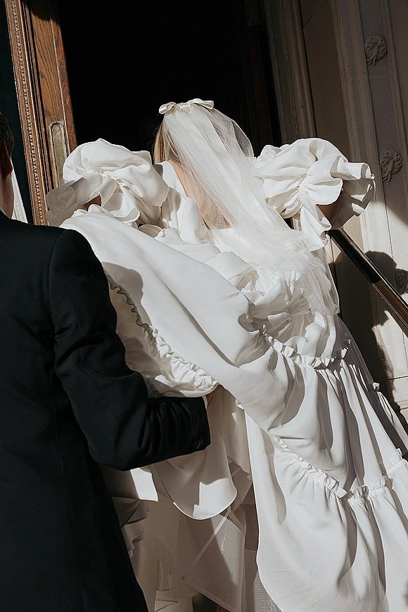 Bride portrait with a bridal veil bow, showing puff sleeve wedding dress and long train on a staircase as groom adjusts her gown in soft light
