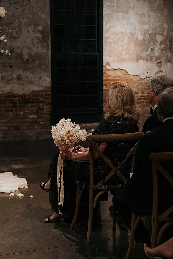 Wedding guest moment as a mother of the bride holds a blush bouquet with white ribbon, seated on cross-back chairs by a rose petal aisle runner