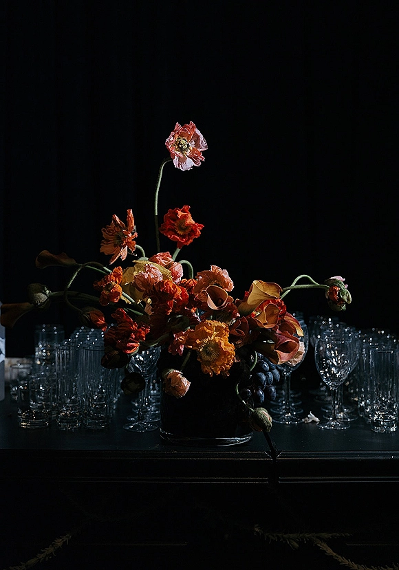 Wedding centerpiece with a moody floral centerpiece of poppies and calla lilies in a clear vase, flanked by grapes and glassware on a dark table.