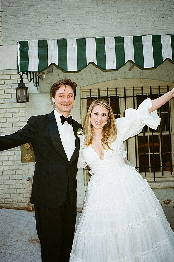 Couple portrait of bride and groom portrait arm in arm, groom in black tuxedo and bride in puff sleeves by a white brick wall under a striped awning