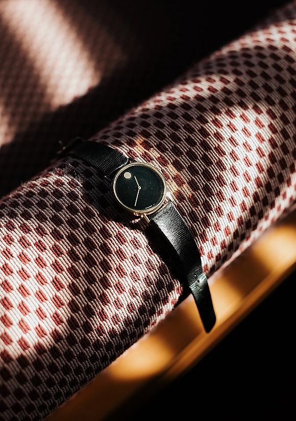 Groom watch with black leather strap and gold case resting on patterned chair upholstery, highlighted by soft sunlight shadows