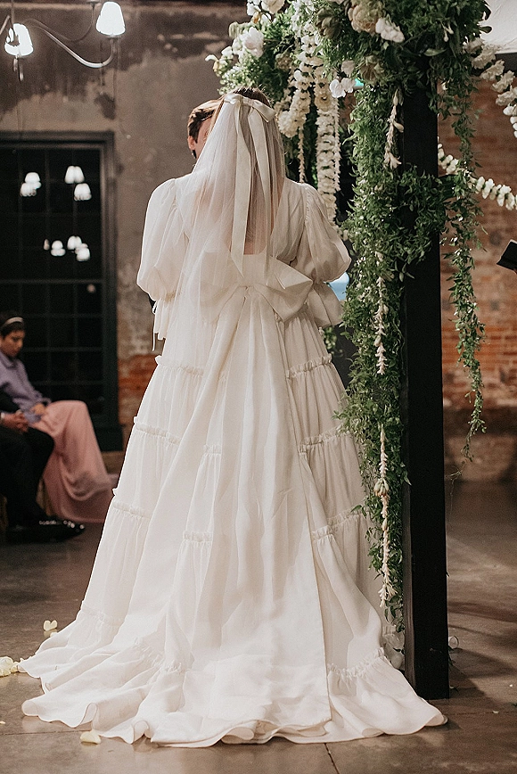 Wedding kiss portrait of bride and groom kissing under a floral arch, bride in veil with oversized bow, guests seated against brick wall backdrop