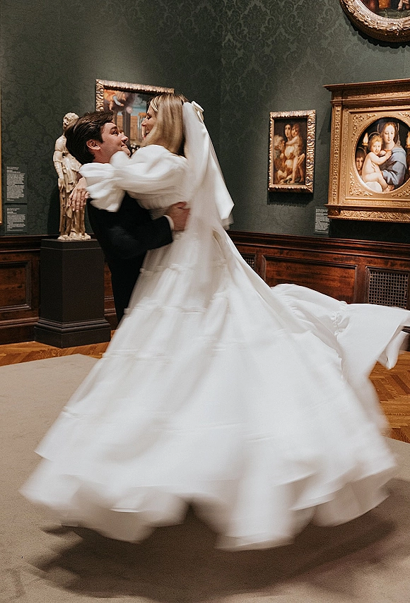 Couple portrait of bride in a long veil and flowing wedding dress with groom in a black suit, dancing in an art gallery with framed paintings