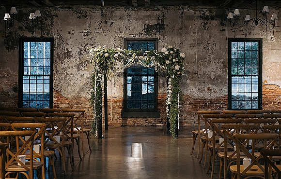 Ceremony setup with an indoor ceremony arch of greenery and white flowers, wooden chairs and chandeliers against an industrial brick wall
