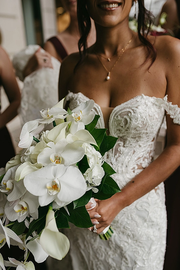 Bridal portrait of a bride holding bouquet of orchids and calla lilies in a strapless lace gown, with bridesmaids blurred in doorway behind