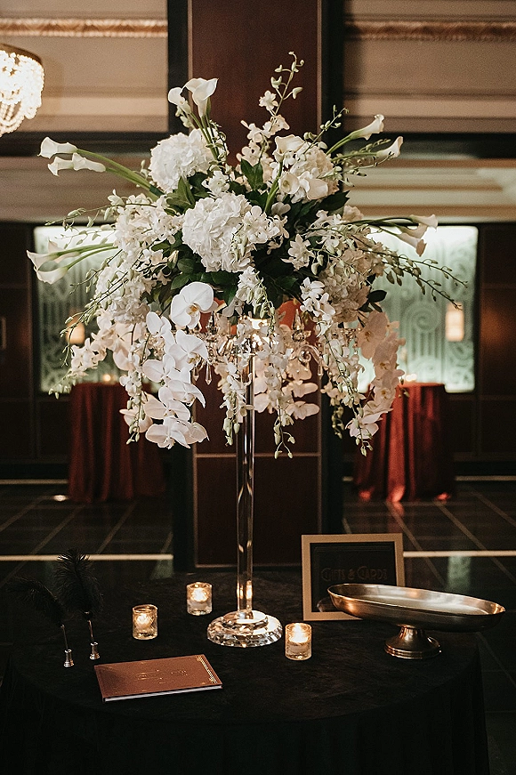 Wedding centerpiece with tall wedding centerpiece in a glass vase, white hydrangeas and orchids, votive candles, guest book under chandelier