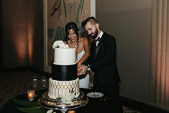 Wedding cake cutting as bride and groom slice a three-tier white cake with flowers, champagne flutes and votive candles on black tablecloth indoors