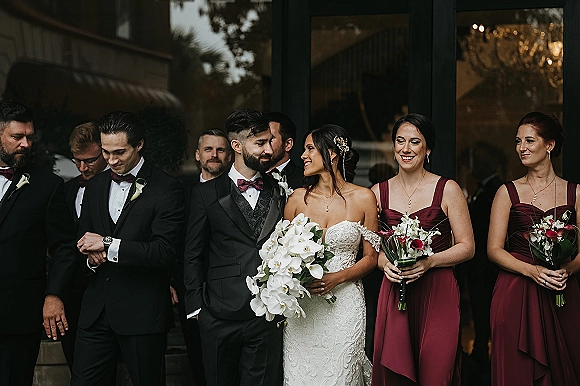 Wedding party portrait with bride and groom with bridal party at glass-door entrance, bride holding cascading orchid bouquet with greenery reflections