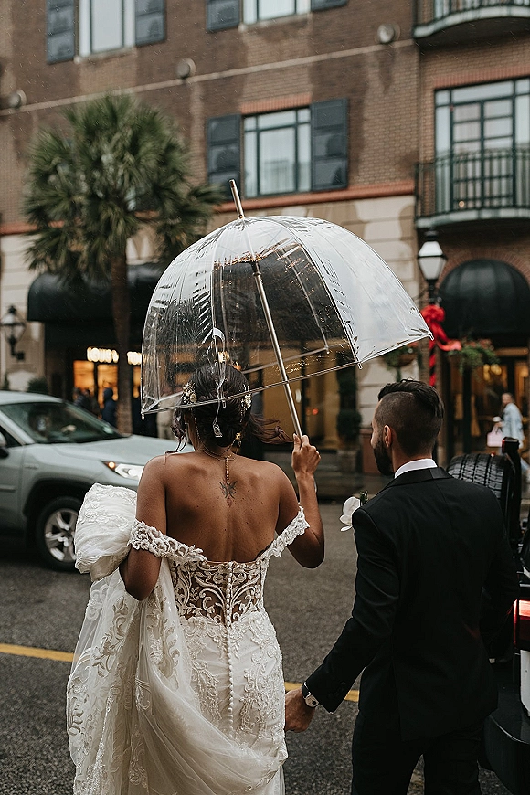 Wedding exit moment with newlyweds holding hands under a clear umbrella, walking toward a wedding car on a rainy city street