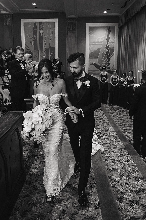 Wedding recessional with bride and groom walking aisle, holding hands as guests clap indoors, bride in strapless lace gown with orchid bouquet