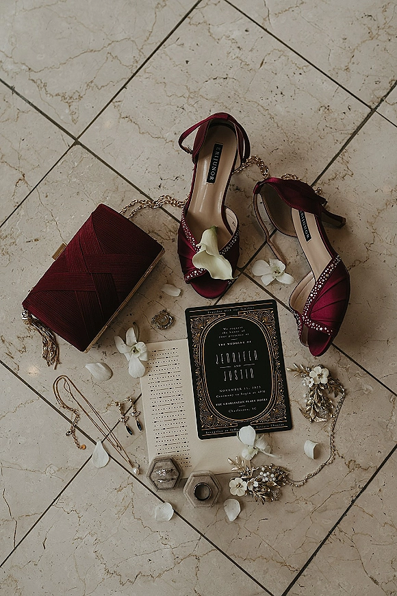 Wedding details flat lay with a wedding invitation flat lay, burgundy heels, clutch, gold jewelry, rings and petals on marble tile floor