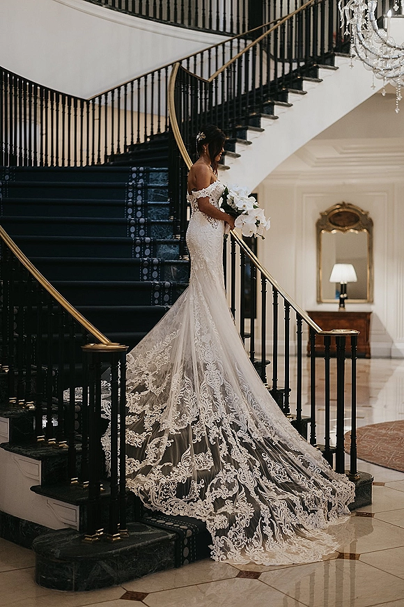 Bridal portrait of bride on staircase in a lace off-the-shoulder gown with long train, holding a white orchid bouquet in a hotel lobby