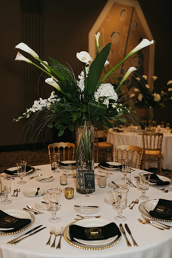 Reception tablescape with tall wedding centerpiece of white calla lilies and greenery, black napkins with gold chargers, in a banquet hall