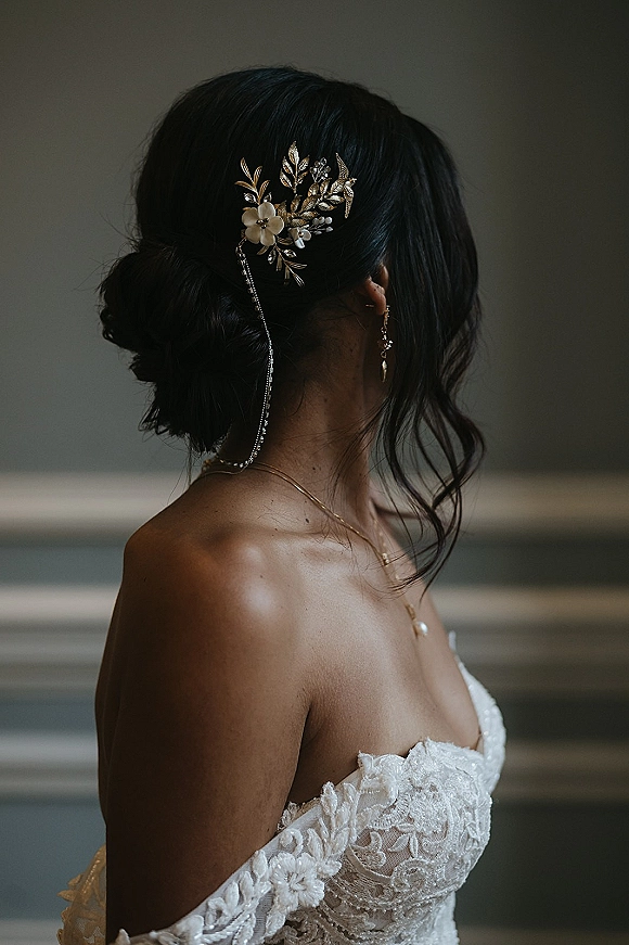 Bridal hair accessory with a gold wedding hair comb and chain pinned into a low bun updo, loose face-framing curls, lace off-shoulder dress indoors