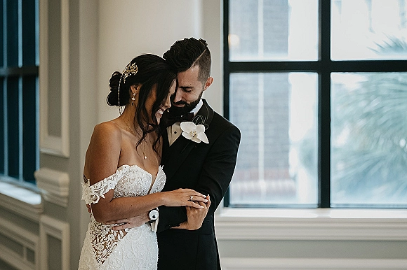 Couple portrait of bride and groom embrace by a large window, forehead touching in soft natural light, lace off-shoulder gown and tuxedo