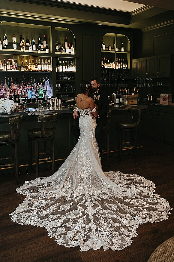 Couple portrait of bride and groom at bar, embracing by a marble counter, her lace train and orchid accents against dark shelves