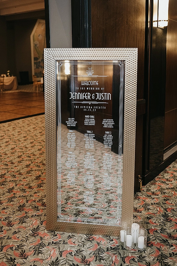 Wedding seating chart mirror sign in a framed mirror with art deco typography and geometric border, flanked by candles in a hallway