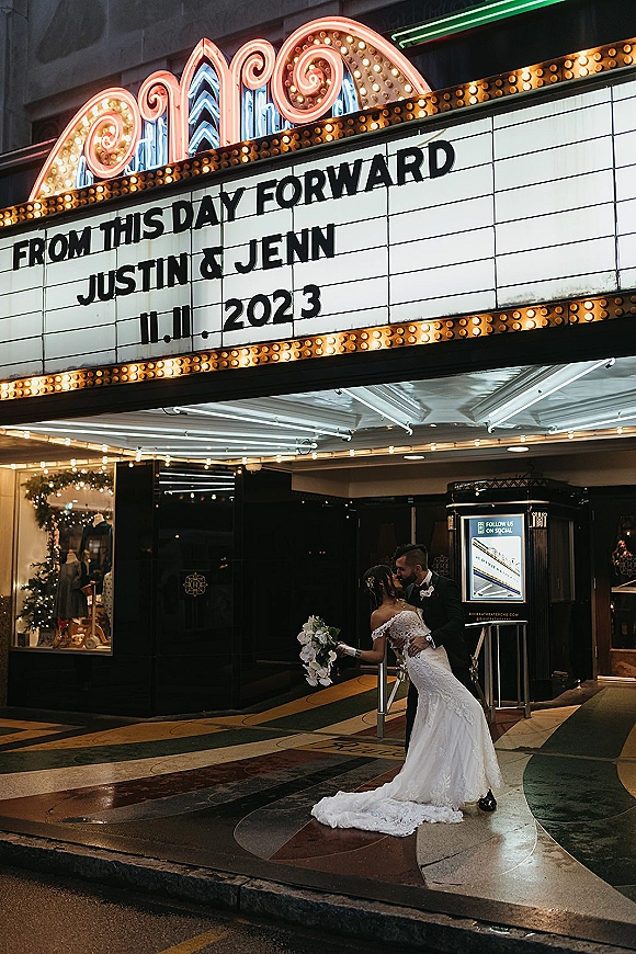 Wedding kiss portrait of bride and groom kissing under a theater marquee, her lace dress and orchid bouquet glowing in neon lights at night