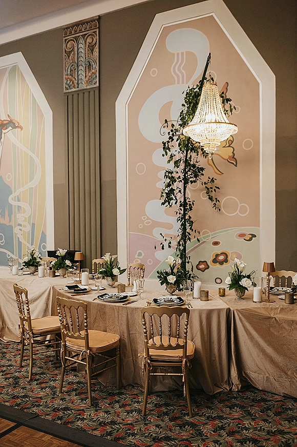Reception tablescape with beige table linens, white floral centerpieces and candles beneath a crystal chandelier, set against a painted wall mural