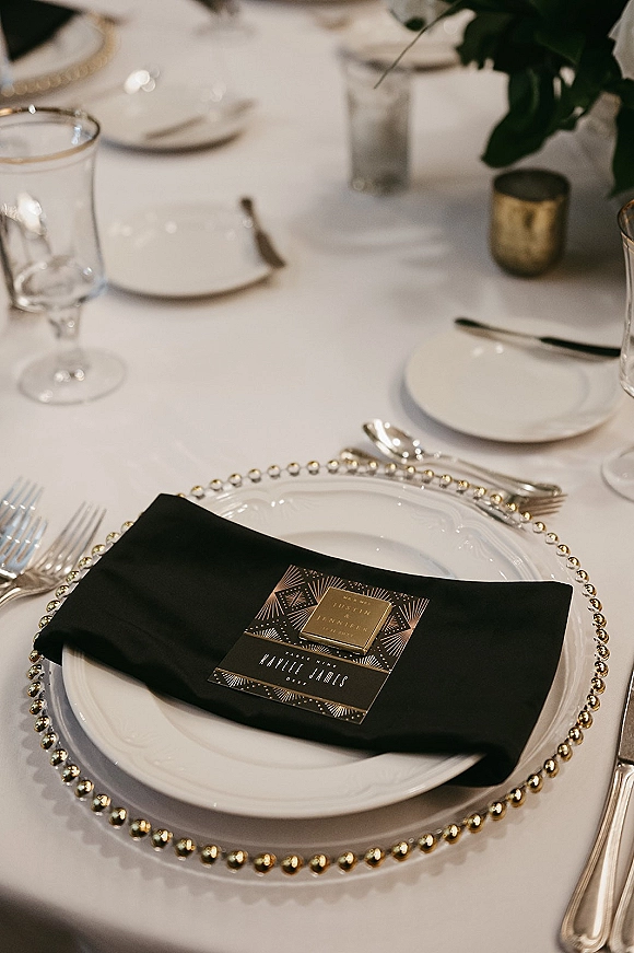 Wedding place setting with a black napkin, white plate on a gold beaded charger, menu and place card, silver flatware and glassware on a white tablecloth