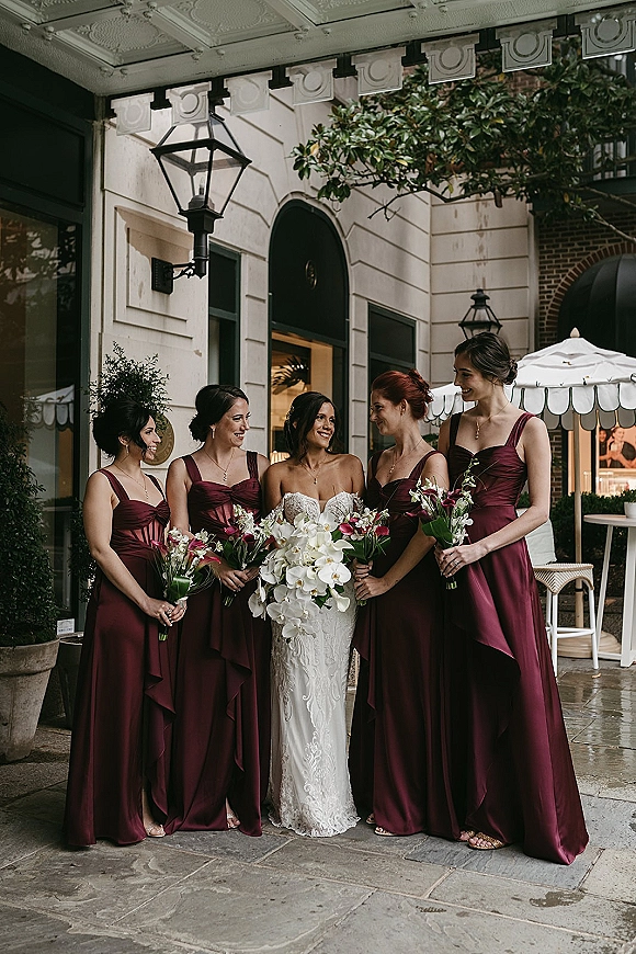 Bride with bridesmaids in burgundy bridesmaid dresses holding bouquets, bride in strapless lace gown with white orchid bouquet at arched entryway