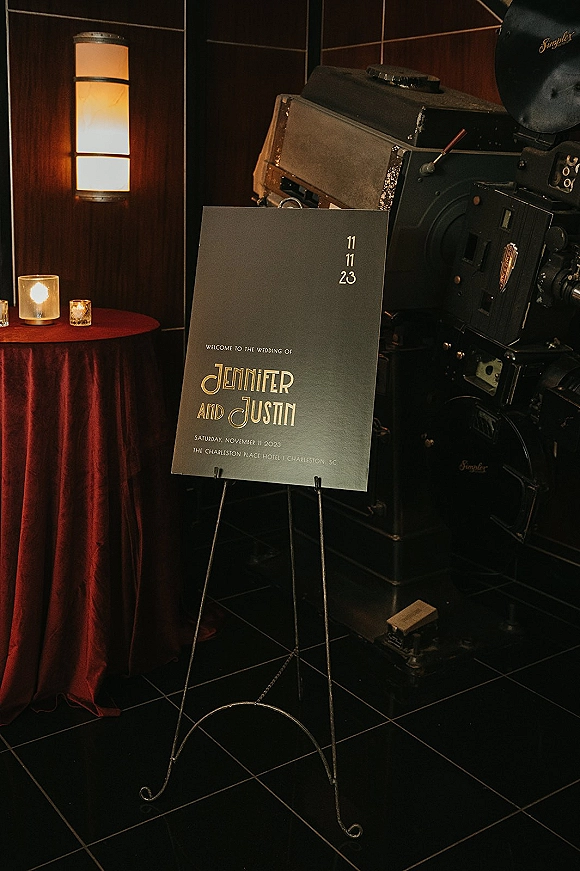Wedding welcome sign on an easel with candles on a linen-draped round table against a dark tiled wall, near a vintage film projector