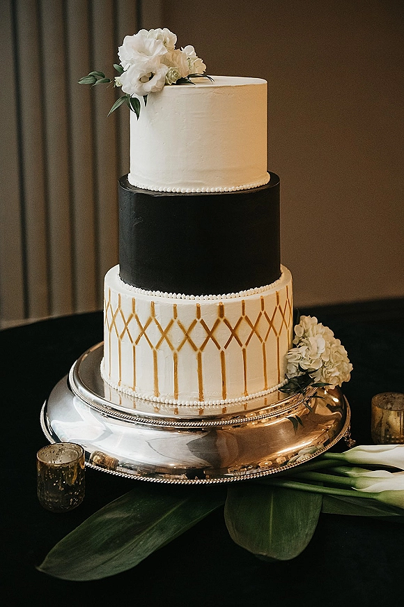 Wedding cake with black and white tiers, gold geometric pattern, and white flowers with greenery on a silver stand by votive candles