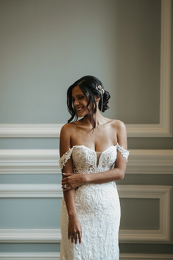 Bridal portrait of a smiling bride in a strapless lace wedding dress, wearing a hair comb and wedding ring against a paneled wall indoors