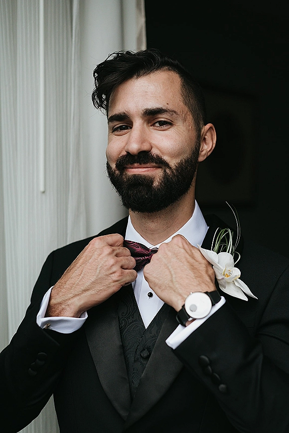 Groom portrait adjusting bow tie in a black tuxedo with patterned waistcoat and white orchid boutonniere in a doorway by a white column