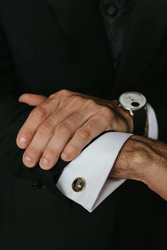 Groom details with groom cufflinks and a wristwatch on a white shirt cuff beneath a black suit jacket against a dark background