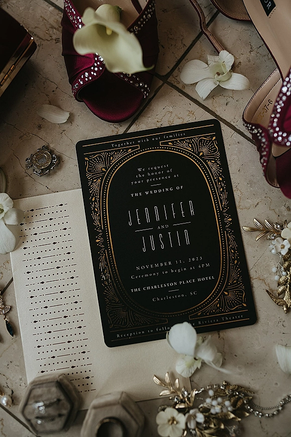 Wedding invitation suite with a black and gold wedding invitation, envelope, burgundy rhinestone heels, orchid petals, and jewelry on stone tile floor
