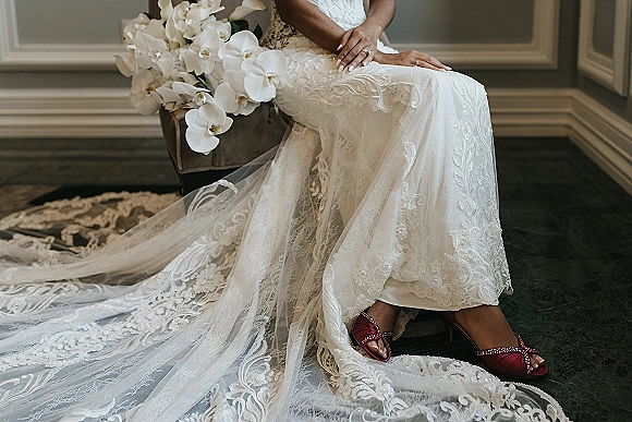 Wedding dress details with lace wedding dress train as the bride sits with veil, orchid bouquet, ring, and red shoes by a paneled wall