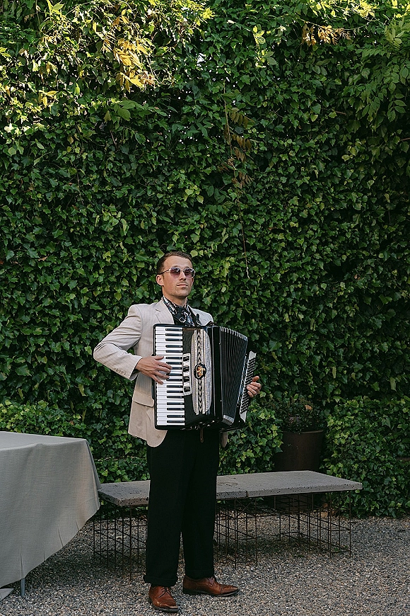 Wedding musician playing an accordion on an outdoor patio, wearing sunglasses and a suit jacket beside an ivy wall backdrop