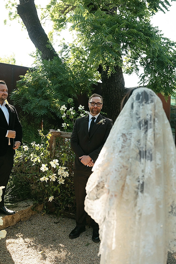 First look moment as bride in lace veil and gown approaches groom in suit and glasses on a gravel garden path with greenery and white flowers