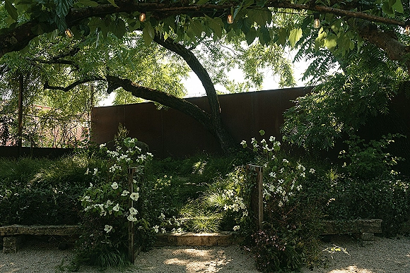 Ceremony aisle design with white floral arrangements and greenery along a gravel path, string lights overhead, framed by garden trees and stone edging