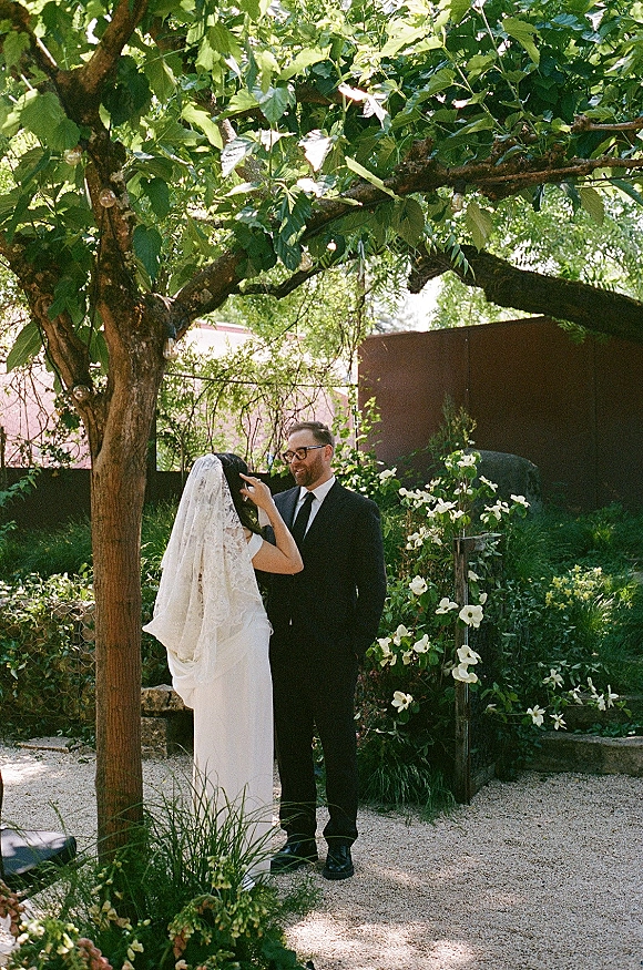 Couple portrait with bride adjusting the groom’s tie beneath garden trees, her lace veil draped as string lights glow over a gravel path