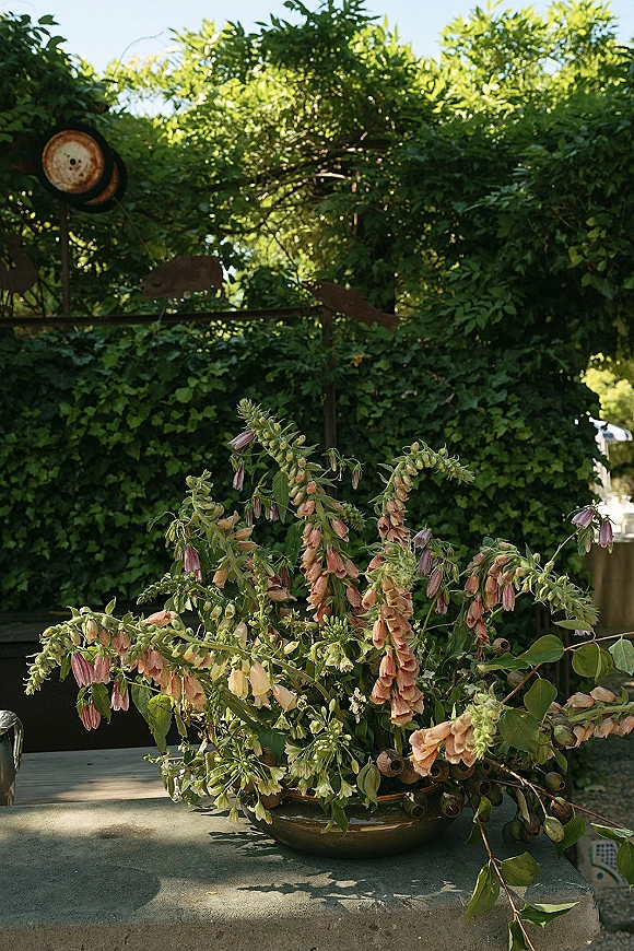 Wedding floral centerpiece in a ceramic bowl with greenery and blush blooms on a stone table, set against garden foliage and rustic metal decor