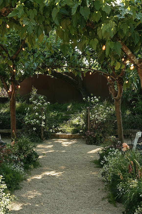 Ceremony aisle design with string lights overhead, ground florals and wooden posts lining a gravel path beneath a tree canopy in a garden