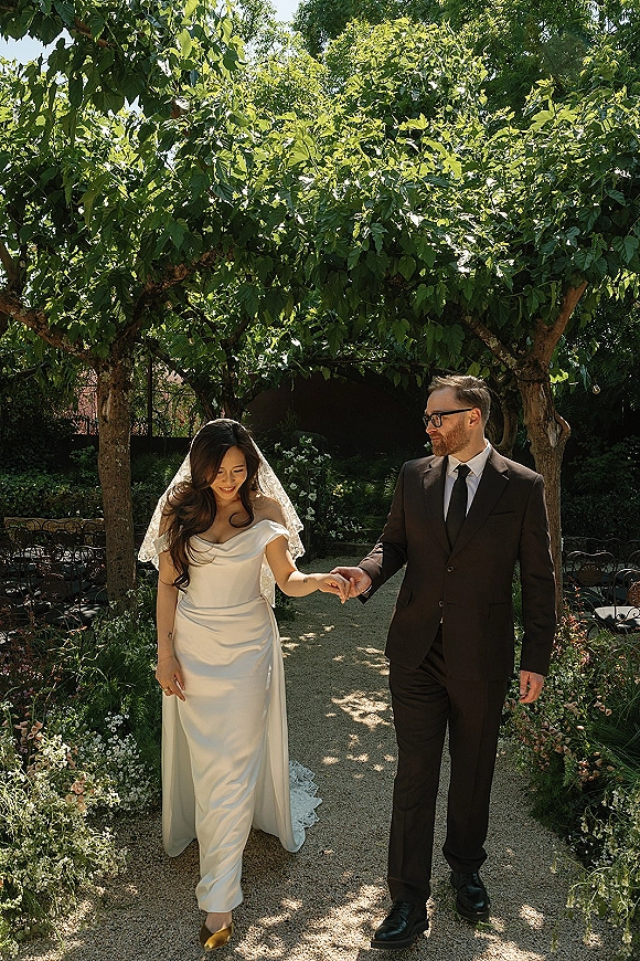 Couple portrait of bride and groom walking hand in hand, bride in a lace veil and off-shoulder dress along a garden path under trees