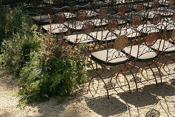 Ceremony seating with wrought iron wedding chairs and cushioned seats, lined with greenery on gravel ground in sunlit shadows